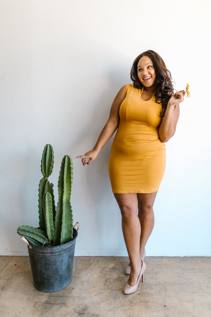 A joyful plus size woman in a mustard dress poses with a cactus against a white wall, indoors.